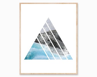 May include: Abstract art print featuring a geometric triangle design. The triangle is divided into sections with shades of blue, gray, and black, evoking a landscape or mountain scene. The artwork is framed in a light-colored wooden frame.
