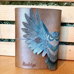 May include: A brown leather journal with an owl design. The owl has blue and teal wings and a silver tree of life charm. The name "Madelyn" is embossed on the front. A blue elastic band keeps the journal closed.