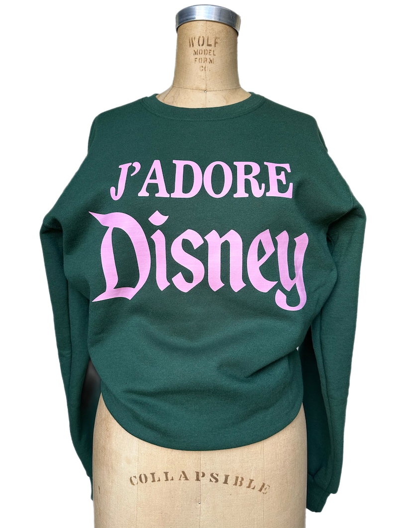 Op de afbeelding: Een donkergroen sweatshirt met de tekst "J'ADORE Disney" in roze letters.