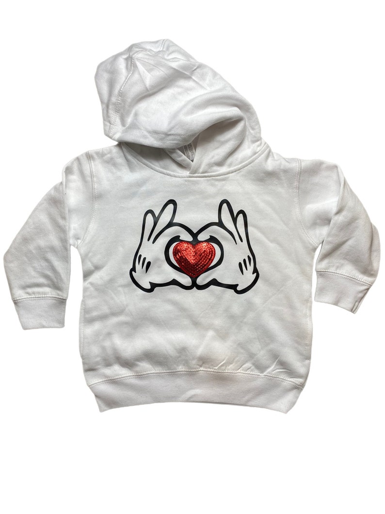 May include: White hooded sweatshirt featuring a graphic of black gloved hands forming a heart shape with a red sequined heart in the center. The sweatshirt has long sleeves and a hood. The design is centered on the front of the garment.