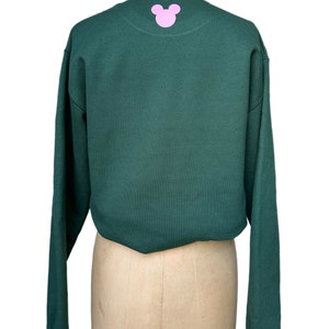 Op de afbeelding: Een donkergroen sweatshirt met lange mouwen en een roze Mickey Mouse-silhouet op de rug.