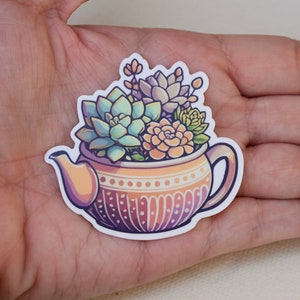 May include: A sticker of a teapot filled with succulents. The teapot is decorated with a brown and white pattern. The succulents are green, pink, and orange.