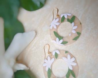 May include: A pair of cream-colored hoop earrings with white flowers and green leaves. The earrings are made of polymer clay and have gold hooks.