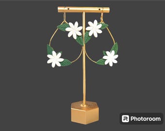 May include: A pair of gold hoop earrings with white flower and green leaf accents. The earrings are displayed on a gold stand.