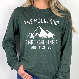 May include: Green long-sleeve shirt with the text "THE MOUNTAINS ARE CALLING AND I MUST GO" in white. The graphic features a mountain range. The shirt is made of a soft material and is suitable for outdoor enthusiasts.