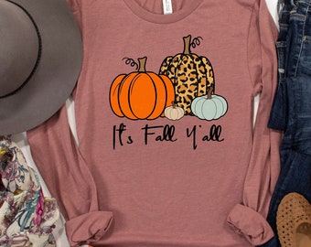 May include: A long-sleeved, mauve t-shirt with a graphic of three pumpkins. The pumpkins are orange, brown with a leopard print, and light blue. The text "It's Fall Y'all" is printed below the pumpkins.