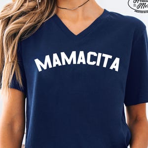 May include: A navy blue V-neck t-shirt featuring the word MAMACITA printed in white across the chest. The upper right corner displays a logo with the text Quality Apparel Healthy Merch Printed in Idaho. The bottom text reads PRINTED IN-HOUSE NOT OUTSOURCED.