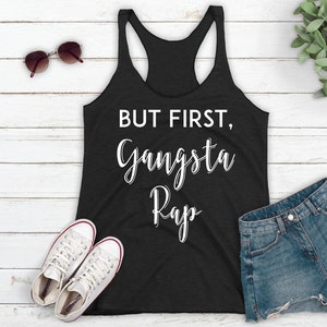 May include: A black tank top with the text "BUT FIRST, Gangsta Rap" in white lettering.