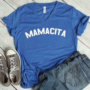 May include: A royal blue v-neck t-shirt with the text "MAMACITA" in white letters. The shirt is laid out on a white wooden surface with a pair of gray canvas shoes and blue denim shorts.