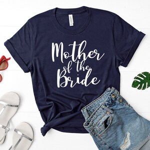 Puede incluir: Una camiseta azul marino con texto blanco que dice "Madre de la novia". La camiseta est&aacute; colocada sobre una superficie blanca con unos pantalones cortos de mezclilla, un par de sandalias plateadas y un llavero de hoja verde.