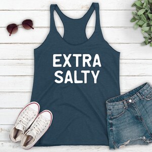 May include: A dark blue tank top with the text "EXTRA SALTY" in white lettering. The tank top is laying on a white wooden surface with a pair of sunglasses, two white sneakers, and a pair of blue denim shorts.