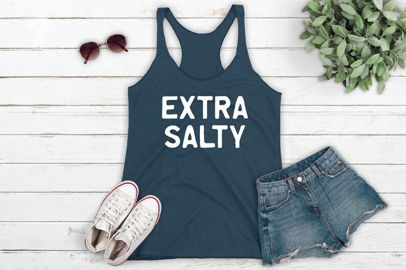 May include: A dark blue tank top with the text "EXTRA SALTY" in white lettering. The tank top is laying on a white wooden surface with a pair of sunglasses, two white sneakers, and a pair of blue denim shorts.