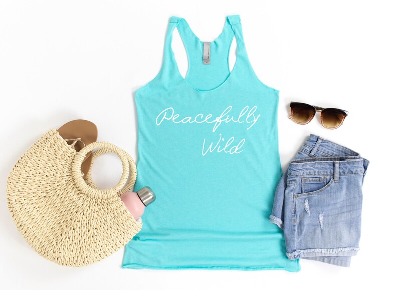 May include: A turquoise tank top with the text "Peacefully Wild" in white lettering. The tank top is laid flat on a white surface with a straw bag, a water bottle, denim shorts, and sunglasses.