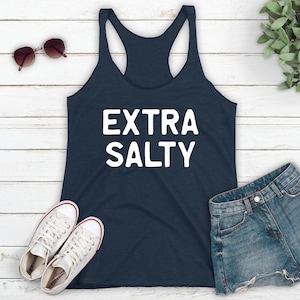 May include: A navy blue tank top with the text "EXTRA SALTY" in white lettering. The tank top is laying on a white wooden surface with a pair of sunglasses, a pair of white sneakers, and a pair of blue denim shorts.
