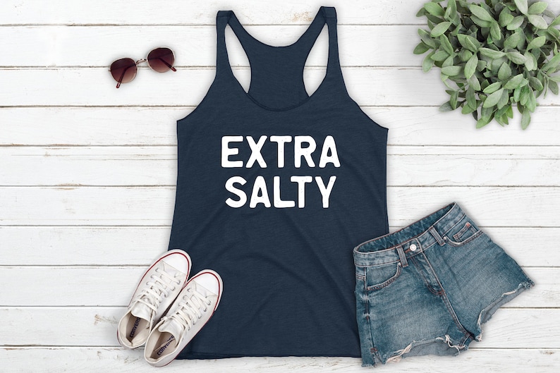 May include: A navy blue tank top with the text "EXTRA SALTY" in white lettering. The tank top is laying on a white wooden surface with a pair of sunglasses, a pair of white sneakers, and a pair of blue denim shorts.