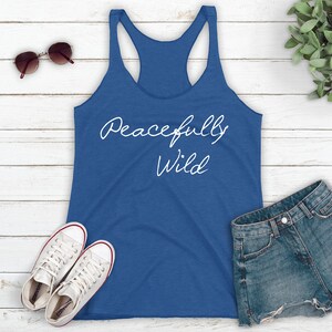 May include: A blue tank top with the text "Peacefully Wild" in white script on the front. The tank top is laying on a white wooden surface with a pair of sunglasses, a pair of white sneakers, and a pair of denim shorts.