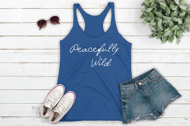 May include: A blue tank top with the text "Peacefully Wild" in white script on the front. The tank top is laying on a white wooden surface with a pair of sunglasses, a pair of white sneakers, and a pair of denim shorts.