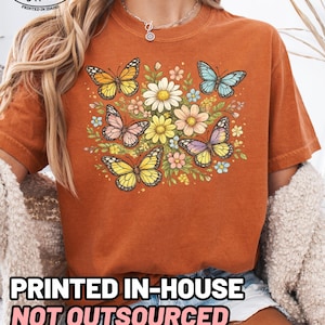 May include: A burnt orange short-sleeve t-shirt featuring a central graphic of four colorful butterflies surrounded by various flowers in shades of yellow, pink, and blue. The shirt has a crew neckline and a relaxed fit.