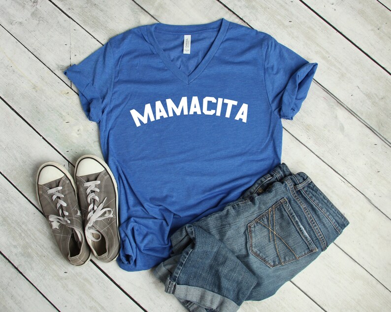 May include: A royal blue v-neck t-shirt with the text "MAMACITA" in white letters. The shirt is laid out on a white wooden surface with a pair of gray canvas shoes and blue denim shorts.