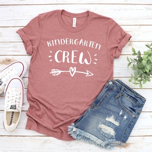 May include: A mauve t-shirt with the text "Kindergarten Crew" in white with a white arrow and heart graphic. The shirt is laid flat on a white wood background with a pair of white sneakers and denim shorts.