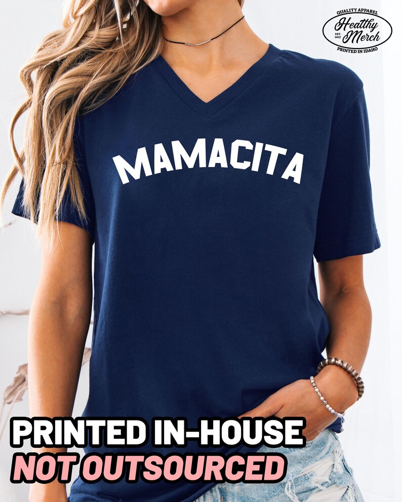 May include: A navy blue V-neck t-shirt featuring the word MAMACITA printed in white across the chest. The upper right corner displays a logo with the text Quality Apparel Healthy Merch Printed in Idaho. The bottom text reads PRINTED IN-HOUSE NOT OUTSOURCED.
