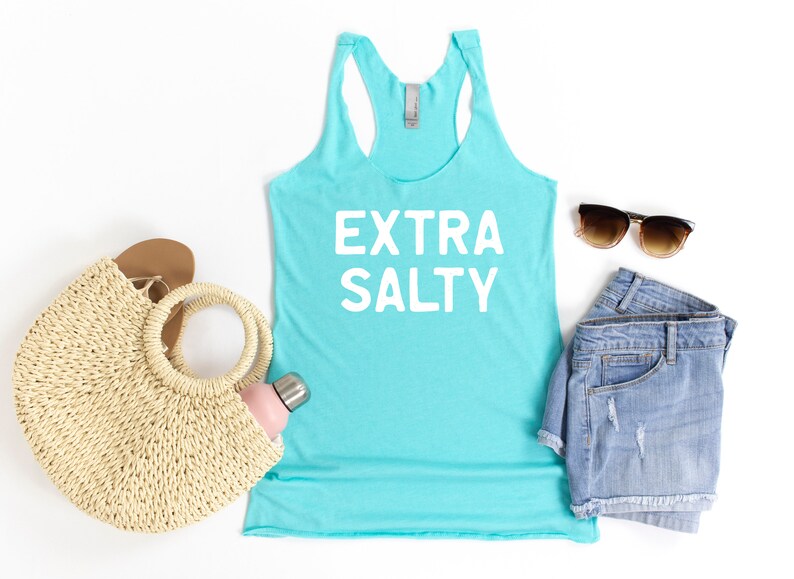 May include: A turquoise blue tank top with the text "EXTRA SALTY" in white. The tank top is styled with denim shorts, sunglasses, a straw bag, and a water bottle.