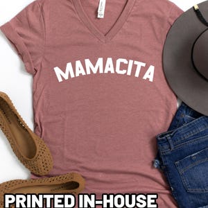 May include: A mauve V-neck t-shirt with the word MAMACITA printed in white across the chest. The shirt is accompanied by a grey wide-brimmed hat, blue denim shorts, and tan woven flat shoes. The shirt features a white neck label.