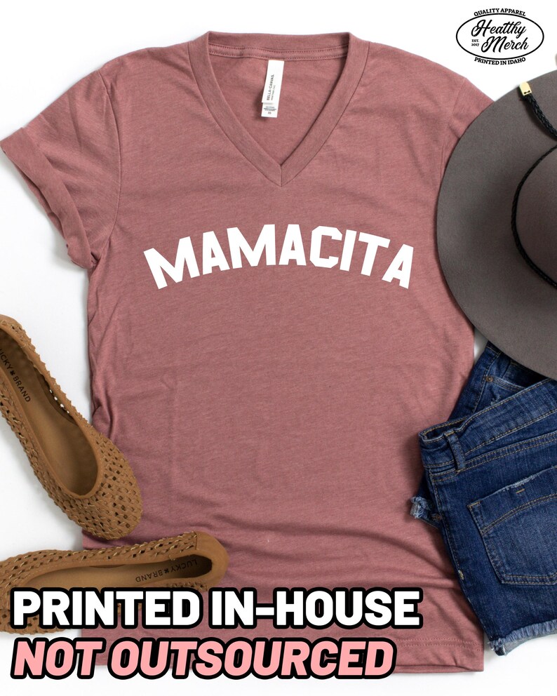 May include: A mauve V-neck t-shirt with the word MAMACITA printed in white across the chest. The shirt is accompanied by a grey wide-brimmed hat, blue denim shorts, and tan woven flat shoes. The shirt features a white neck label.