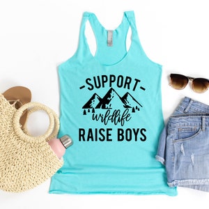 May include: A turquoise tank top with a black graphic that says "Support wildlife raise boys" with a mountain silhouette. The tank top is laid flat on a white background with a straw bag, a water bottle, a pair of denim shorts, and sunglasses.
