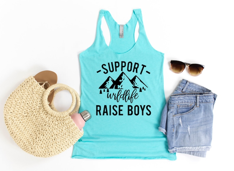 May include: A turquoise tank top with a black graphic that says "Support wildlife raise boys" with a mountain silhouette. The tank top is laid flat on a white background with a straw bag, a water bottle, a pair of denim shorts, and sunglasses.