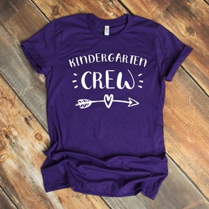 May include: Purple t-shirt with white text that reads "Kindergarten Crew" with an arrow and heart graphic.