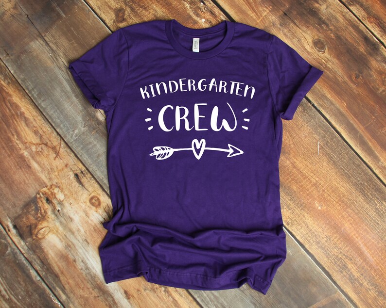 May include: Purple t-shirt with white text that reads "Kindergarten Crew" with an arrow and heart graphic.