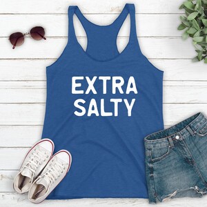 May include: A blue tank top with the text "EXTRA SALTY" in white lettering. The tank top is laying on a white wooden surface with a pair of sunglasses, a pair of white sneakers, and a pair of denim shorts.