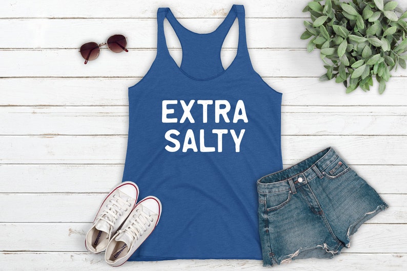 May include: A blue tank top with the text "EXTRA SALTY" in white lettering. The tank top is laying on a white wooden surface with a pair of sunglasses, a pair of white sneakers, and a pair of denim shorts.