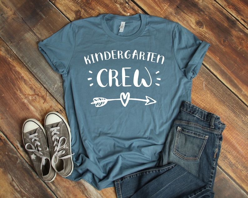 May include: A teal blue t-shirt with the text "Kindergarten Crew" in white with a white arrow and heart design. The shirt is laying on a wood surface with a pair of gray sneakers and blue jeans.