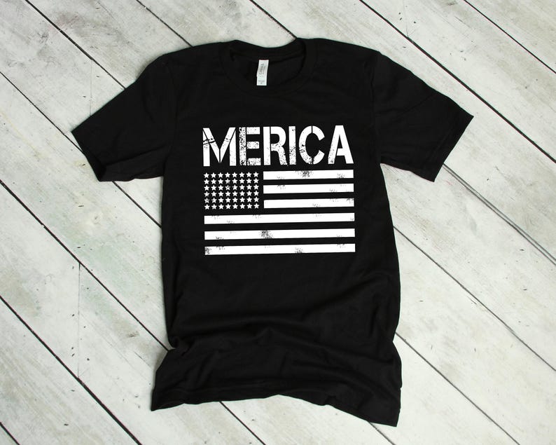 May include: A black short-sleeved t-shirt featuring the white text MERICA printed above a stylized, distressed white graphic of the American flag. The flag design consists of a rectangular field of stars and horizontal stripes in a worn-in style.