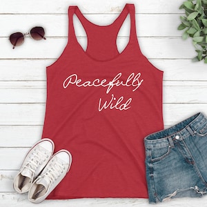 May include: Red tank top with the text "Peacefully Wild" in white script on a white wood background. The tank top is laying flat with a pair of white sneakers and a pair of denim shorts beside it.