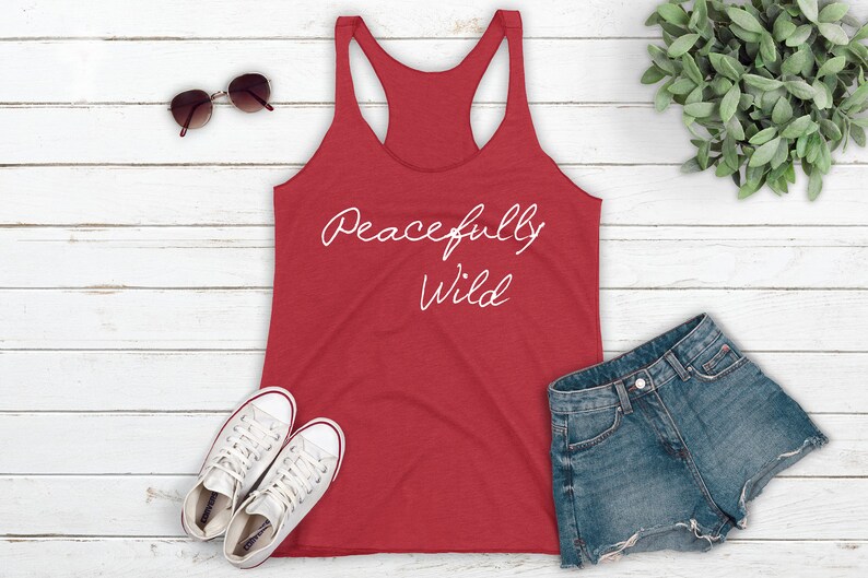 May include: Red tank top with the text "Peacefully Wild" in white script on a white wood background. The tank top is laying flat with a pair of white sneakers and a pair of denim shorts beside it.