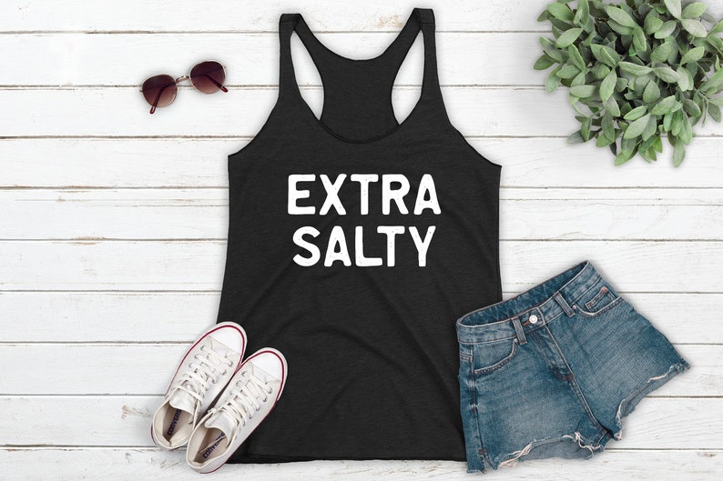 May include: A black tank top with the text "EXTRA SALTY" in white lettering. The tank top is laying on a white wooden surface with a pair of sunglasses, a pair of white sneakers, a pair of denim shorts, and a potted plant.