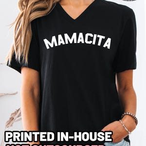 May include: A black V-neck t-shirt featuring the word MAMACITA printed in white capital letters across the chest. The shirt includes a small logo in the top right corner that reads Quality Apparel Healthy Merch Est 2017 Printed in Idaho.