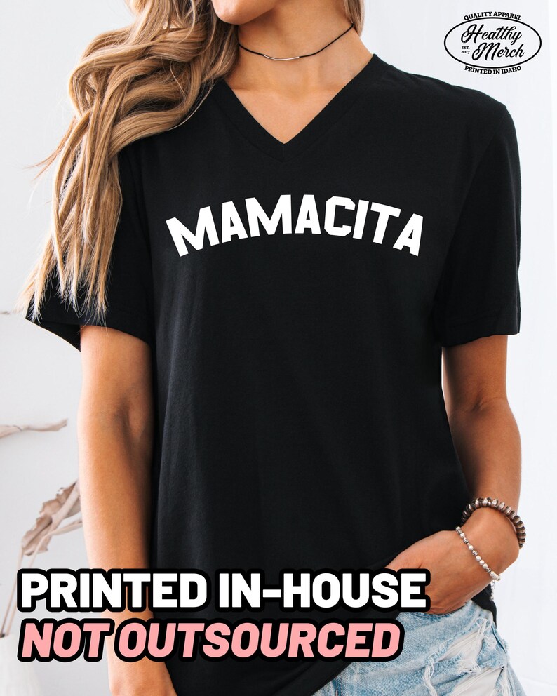 May include: A black V-neck t-shirt featuring the word MAMACITA printed in white capital letters across the chest. The shirt includes a small logo in the top right corner that reads Quality Apparel Healthy Merch Est 2017 Printed in Idaho.