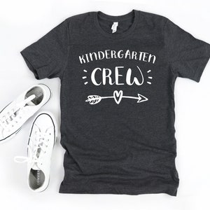 May include: A dark gray t-shirt with the text "Kindergarten Crew" in white lettering. The text is surrounded by white dots and an arrow with a heart at the center.
