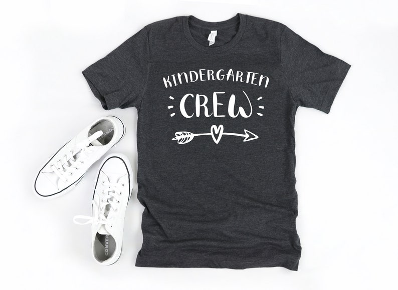 May include: A dark gray t-shirt with the text "Kindergarten Crew" in white lettering. The text is surrounded by white dots and an arrow with a heart at the center.