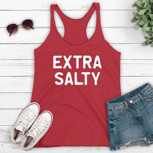 May include: Red tank top with the text "EXTRA SALTY" in white lettering. The tank top is laying on a white wooden surface with a pair of white sneakers, a pair of sunglasses, and a pair of denim shorts.