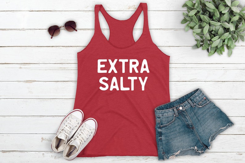 May include: Red tank top with the text "EXTRA SALTY" in white lettering. The tank top is laying on a white wooden surface with a pair of white sneakers, a pair of sunglasses, and a pair of denim shorts.