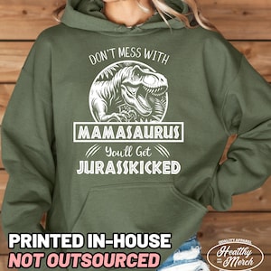 May include: Olive green hooded sweatshirt with a white graphic of a Tyrannosaurus Rex and the text "DON'T MESS WITH MAMASAURUS You'll Get JURASSKICKED." The bottom of the sweatshirt has the text "PRINTED IN-HOUSE NOT OUTSOURCED."