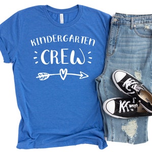 May include: A royal blue t-shirt with the text "Kindergarten Crew" in white lettering. The text is surrounded by small white dots and an arrow with a heart in the middle.