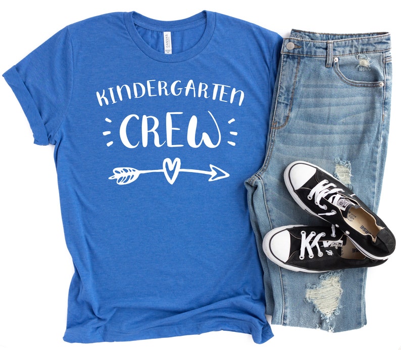May include: A royal blue t-shirt with the text "Kindergarten Crew" in white lettering. The text is surrounded by small white dots and an arrow with a heart in the middle.