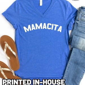 May include: A blue V-neck t-shirt with the word MAMACITA printed in white across the chest. The shirt is paired with blue denim jeans and tan flip-flops. A logo in the top right corner reads Quality Apparel Healthy Merch Printed in Idaho.