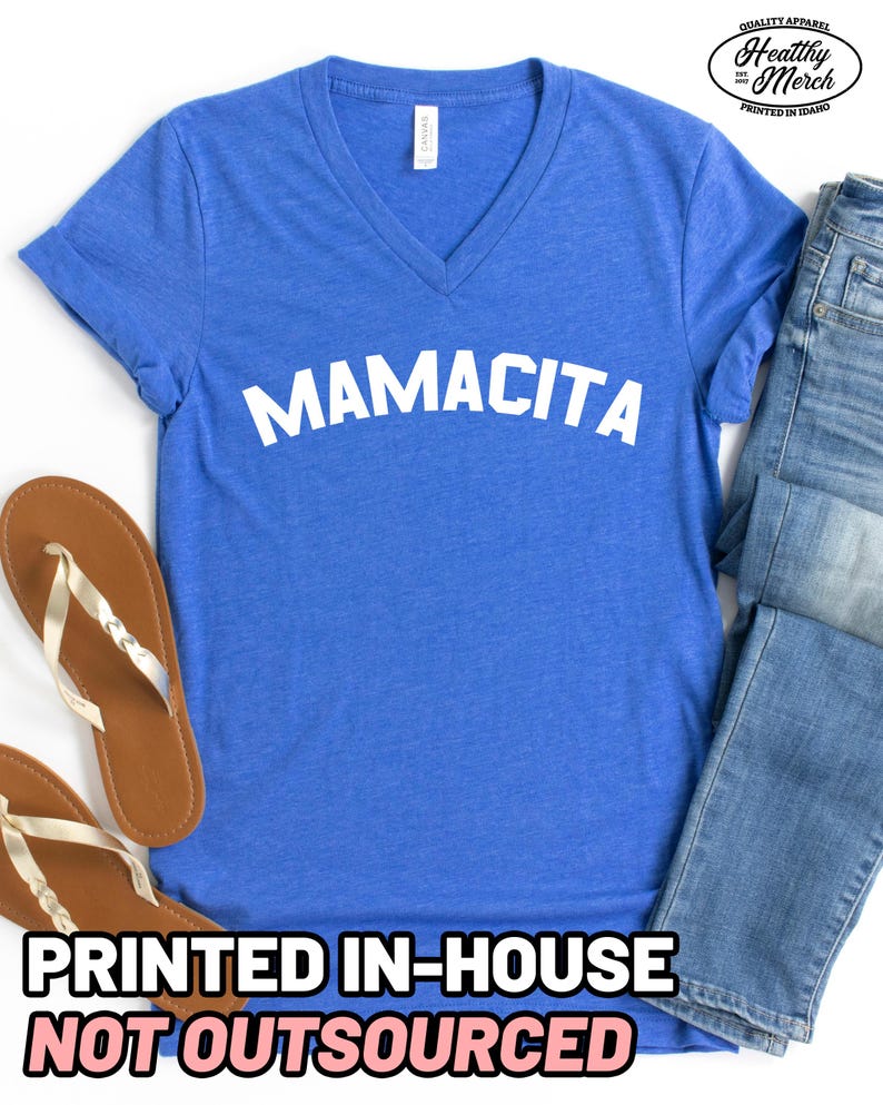 May include: A blue V-neck t-shirt with the word MAMACITA printed in white across the chest. The shirt is paired with blue denim jeans and tan flip-flops. A logo in the top right corner reads Quality Apparel Healthy Merch Printed in Idaho.